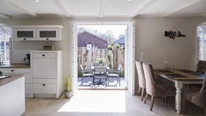 A kitchen with white cabinetry, a dining area with a table and chairs, and a view of a backyard with a wooden fence and a house. - Holiday apartment in Büsum with swimming pool (Büsum)