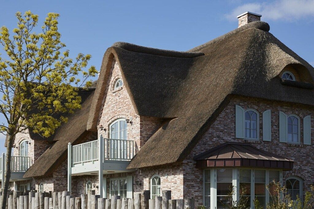 Une maison traditionnelle à toit de chaume, dotée d’une façade en briques, de nombreuses fenêtres et d’une clôture en bois.