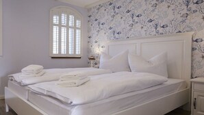 A double bed with white bedding and pillows, a nightstand with a lamp, and a floral wallpaper pattern. - Holiday apartment in Büsum with swimming pool (Büsum)