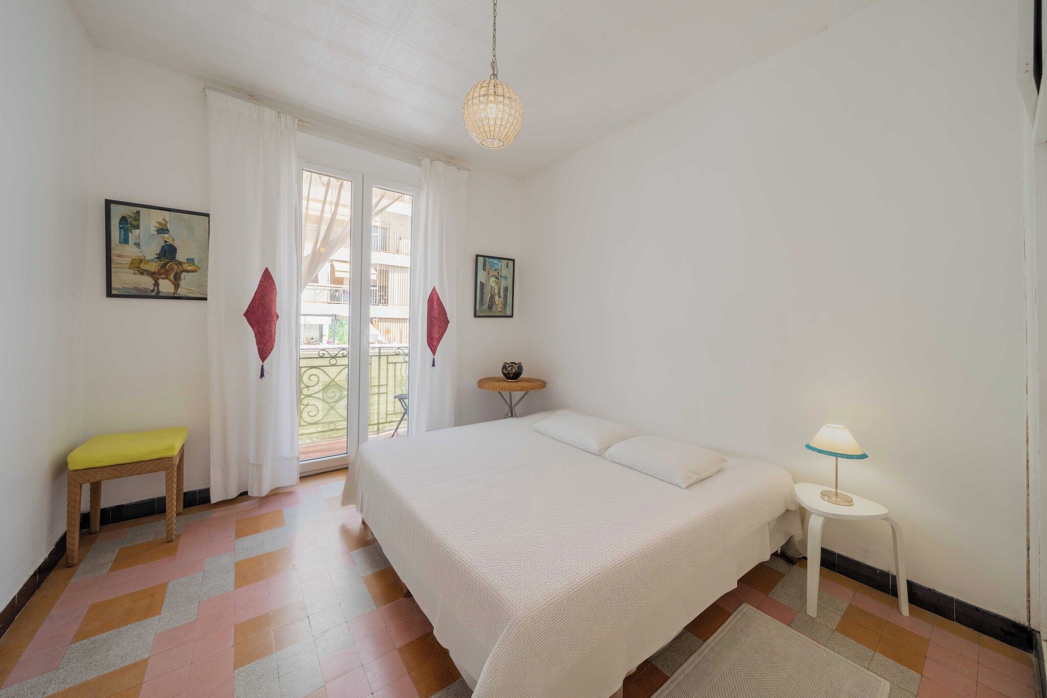 A bedroom with a bed, a small table with a lamp, a chair, and two paintings on the wall.