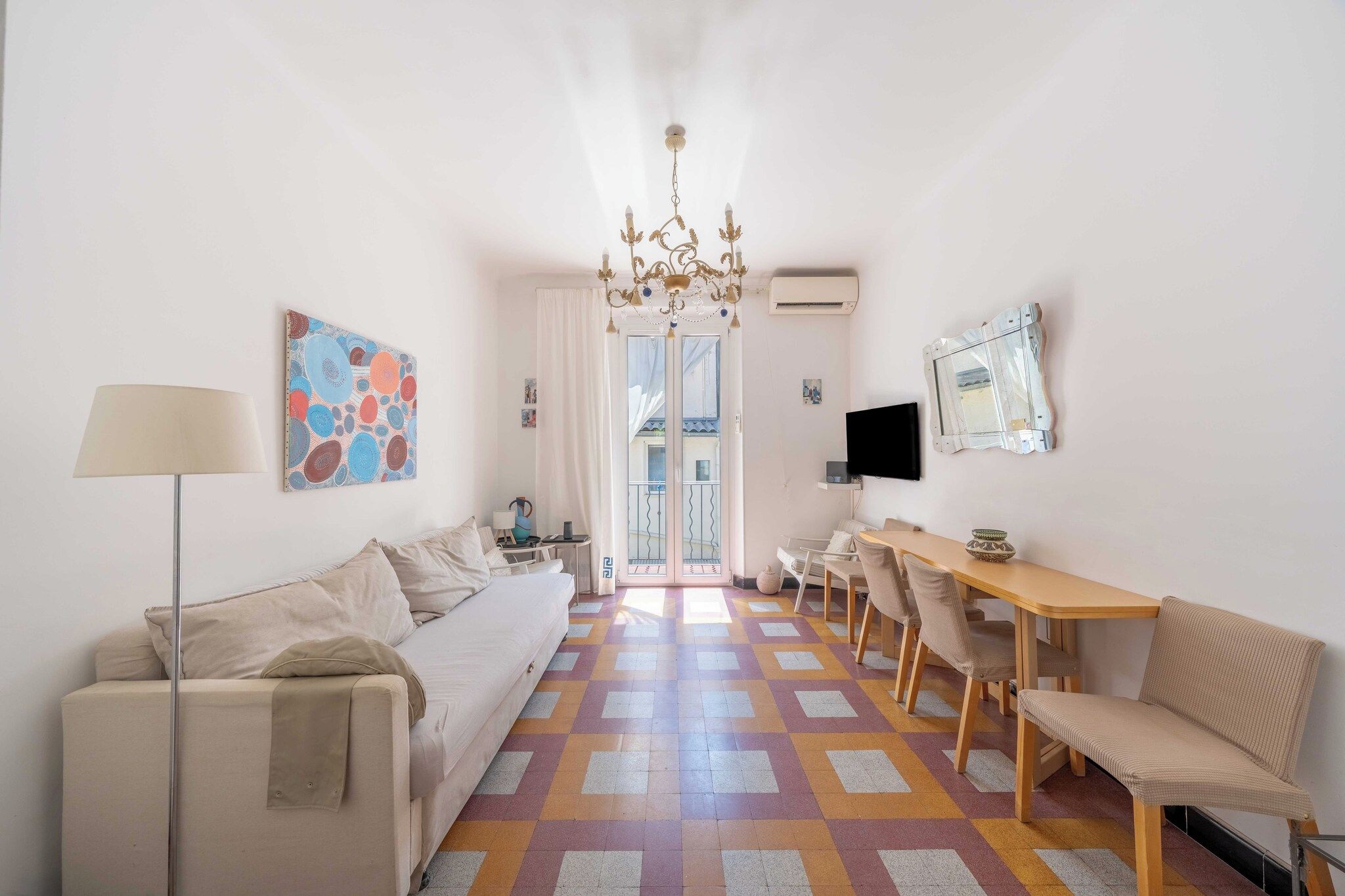 A modern living room with a sofa, a dining table with chairs, a chandelier, and a colorful floor pattern.