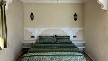 A bedroom with a herringbone-patterned wooden floor, a bed with green patterned bedding, a geometric pendant light, and ornate wall molding.