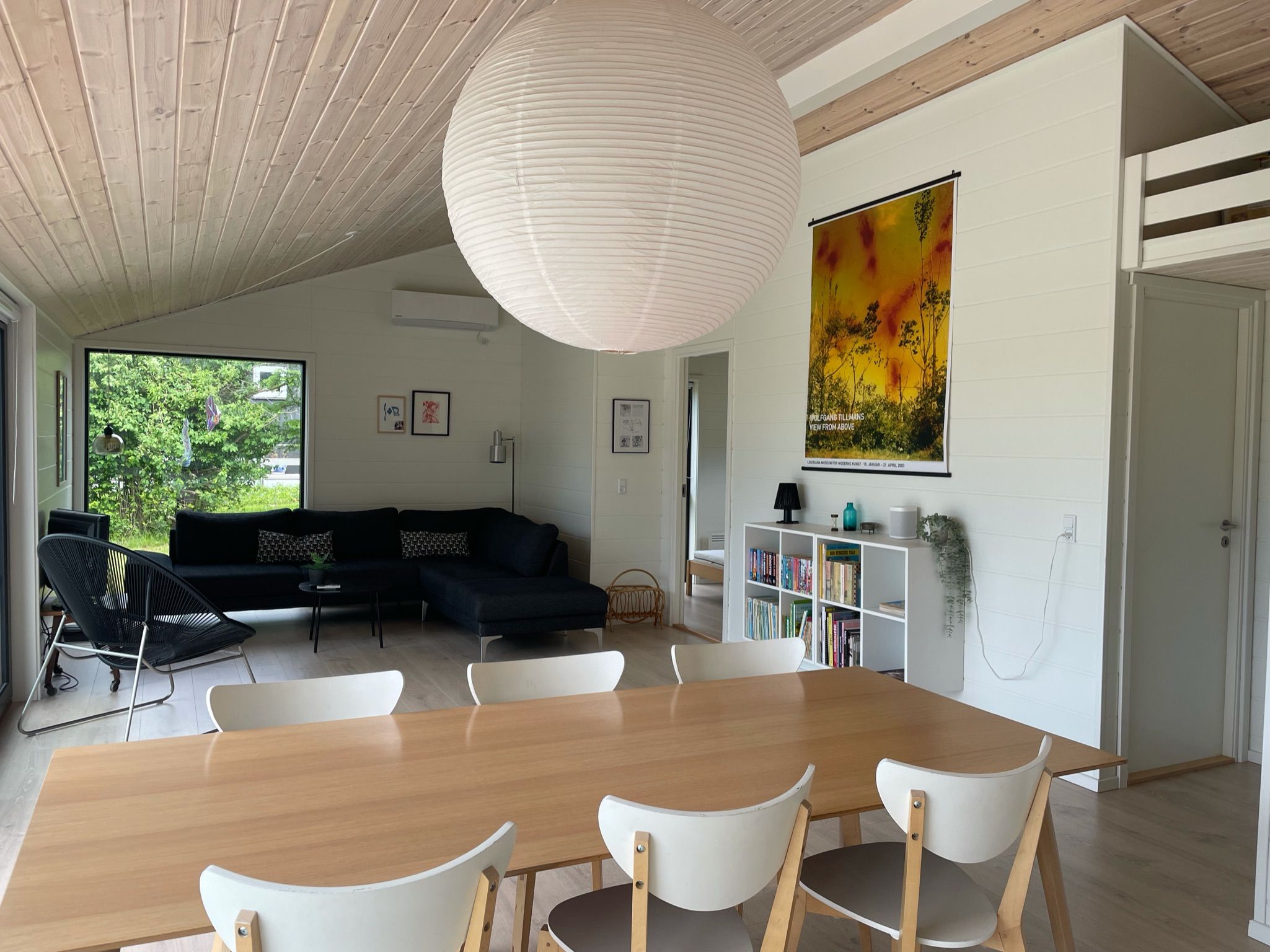 A modern living room with a wooden dining table, white chairs, a black sofa, and a large painting on the wall.