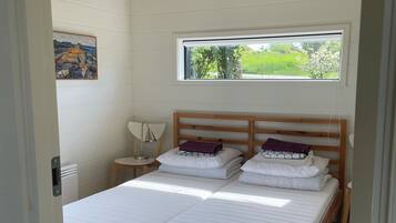 A bedroom with a wooden bed, a bedside table, a lamp, and a window with a view of greenery.