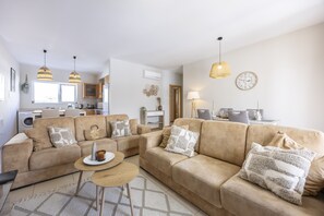 A modern living room with a beige sofa, a coffee table, and a dining area with a chandelier. - Casa do Mar Aromas Condomínio by CoolHouses (Burgau)
