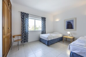 A bedroom with two beds, a chair, a bedside table with a lamp, and a window with patterned curtains. - CoolHouses Algarve Luz | Ramalhete T2 (Luz)