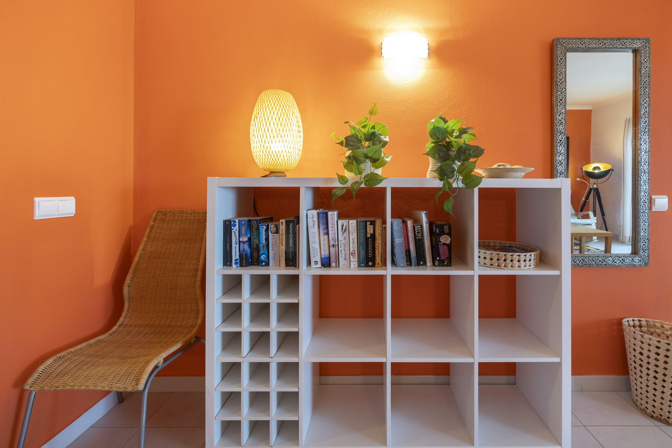 A room with an orange wall, a white bookshelf with books, a wicker chair, a floor lamp, and a mirror with a decorative frame.