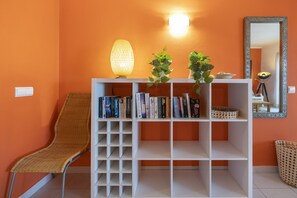 Ein Zimmer mit orangefarbener Wand, einem weißen Bücherregal mit Büchern, einem Korbstuhl, einer Stehlampe und einem Spiegel mit dekorativem Rahmen.