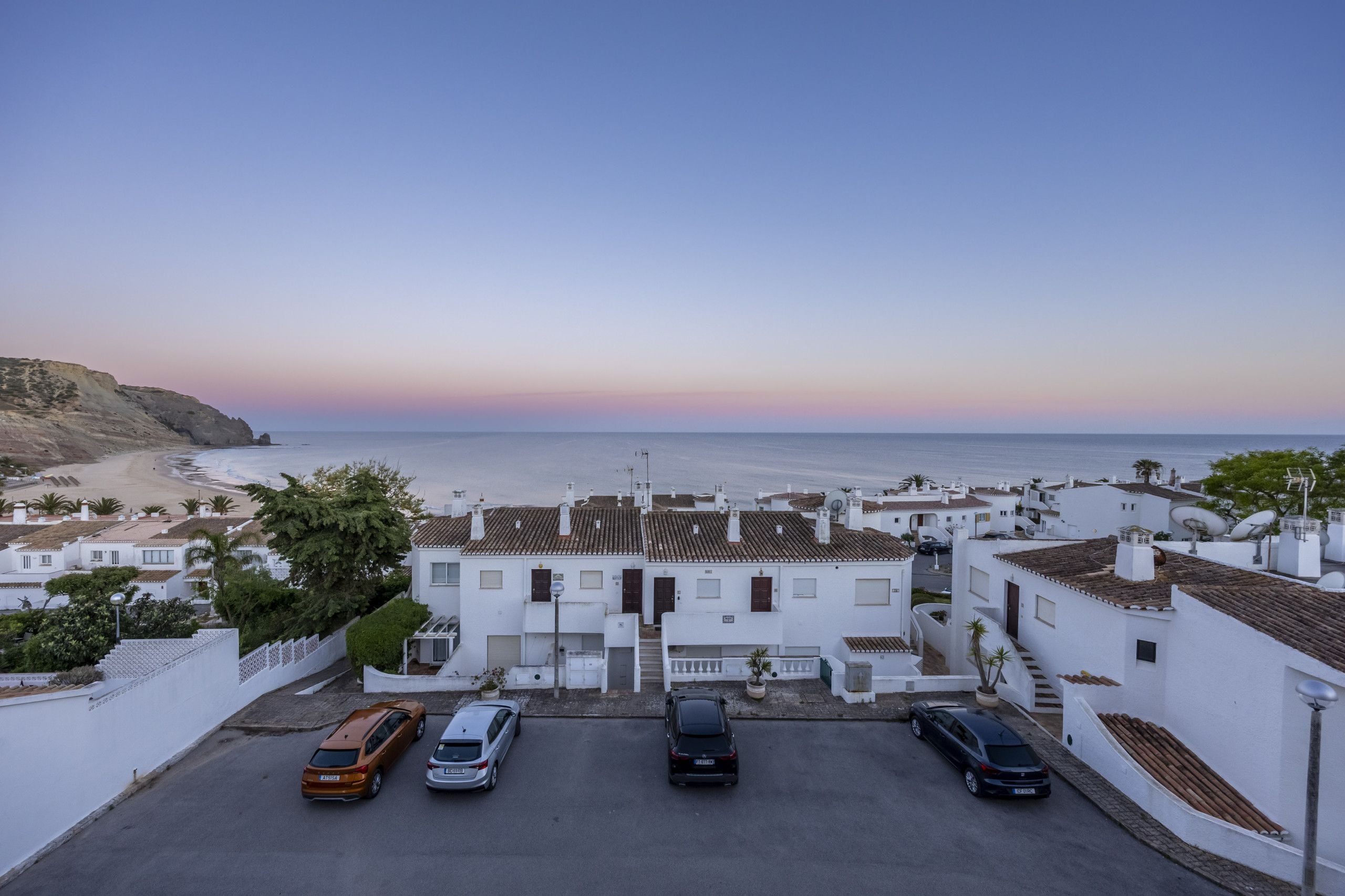 Una località costiera con edifici bianchi, un'area di parcheggio con auto e vista sul mare.