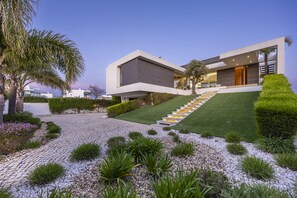 A modern two-story house with a paved driveway, a staircase leading to the entrance, and a well-maintained garden with various plants and a lawn. - Vila Camilo Ponta do Farol by CoolHouses (Lagos)