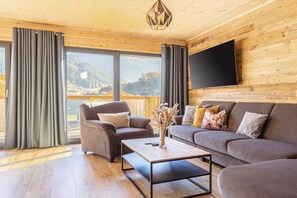 A modern living room with a sofa, armchair, and coffee table, large windows with curtains, and a flat-screen TV on the wall. - Cabin in Steinach am Brenner with sauna (Steinach am Brenner)