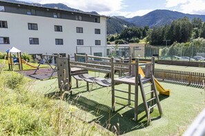 A playground with a slide and climbing structures, surrounded by a building with multiple windows, set against a backdrop of mountains and trees.