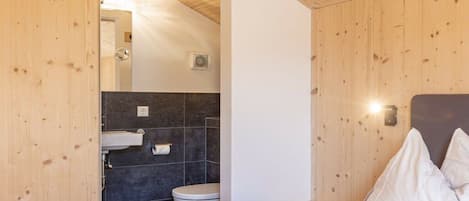 A wooden-paneled room with a bed, white pillows, a bedside table, and a bathroom with a toilet and sink.