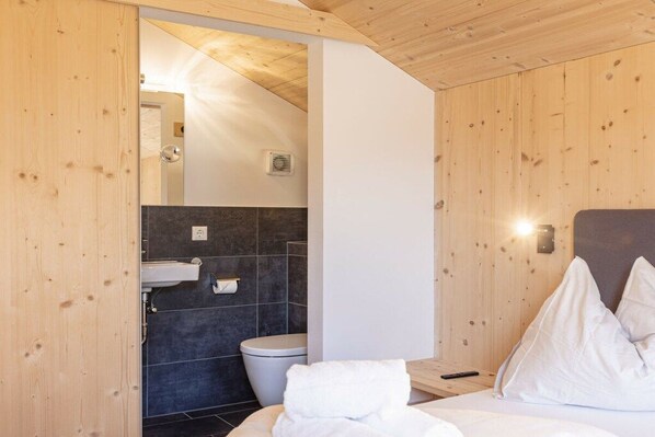 Una habitación con paneles de madera, una cama, almohadas blancas, una mesita de noche y un baño con inodoro y lavamanos.