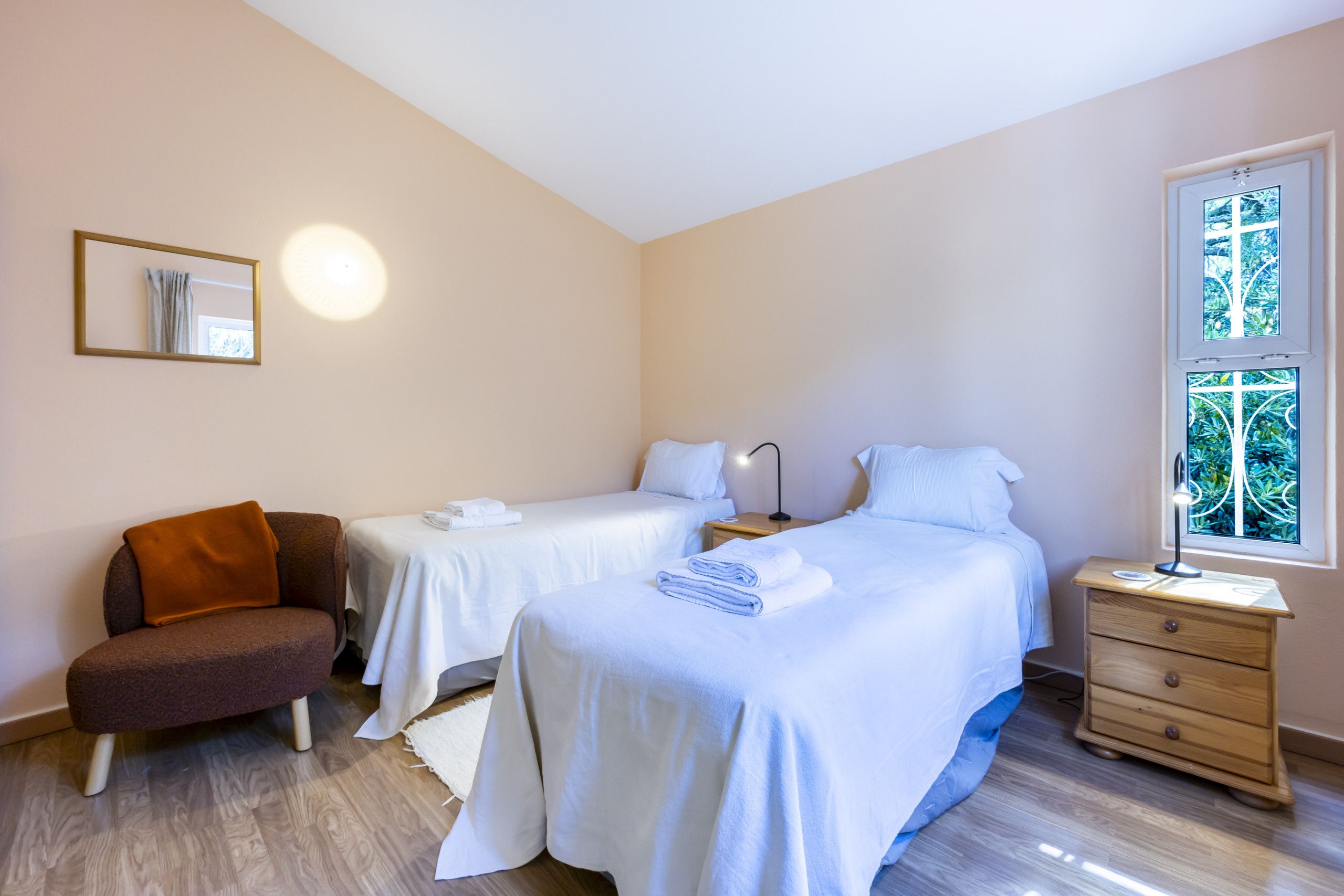 A hotel room with two beds, a chair, a bedside table with a lamp, a mirror, and a window with a view of greenery.