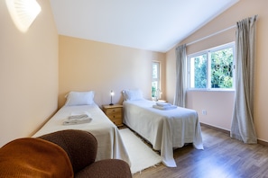 A bedroom with two single beds, a nightstand with a lamp, a window with curtains, and a view of greenery outside. - Quinta da Belavista 55 by CoolHouses (Luz)