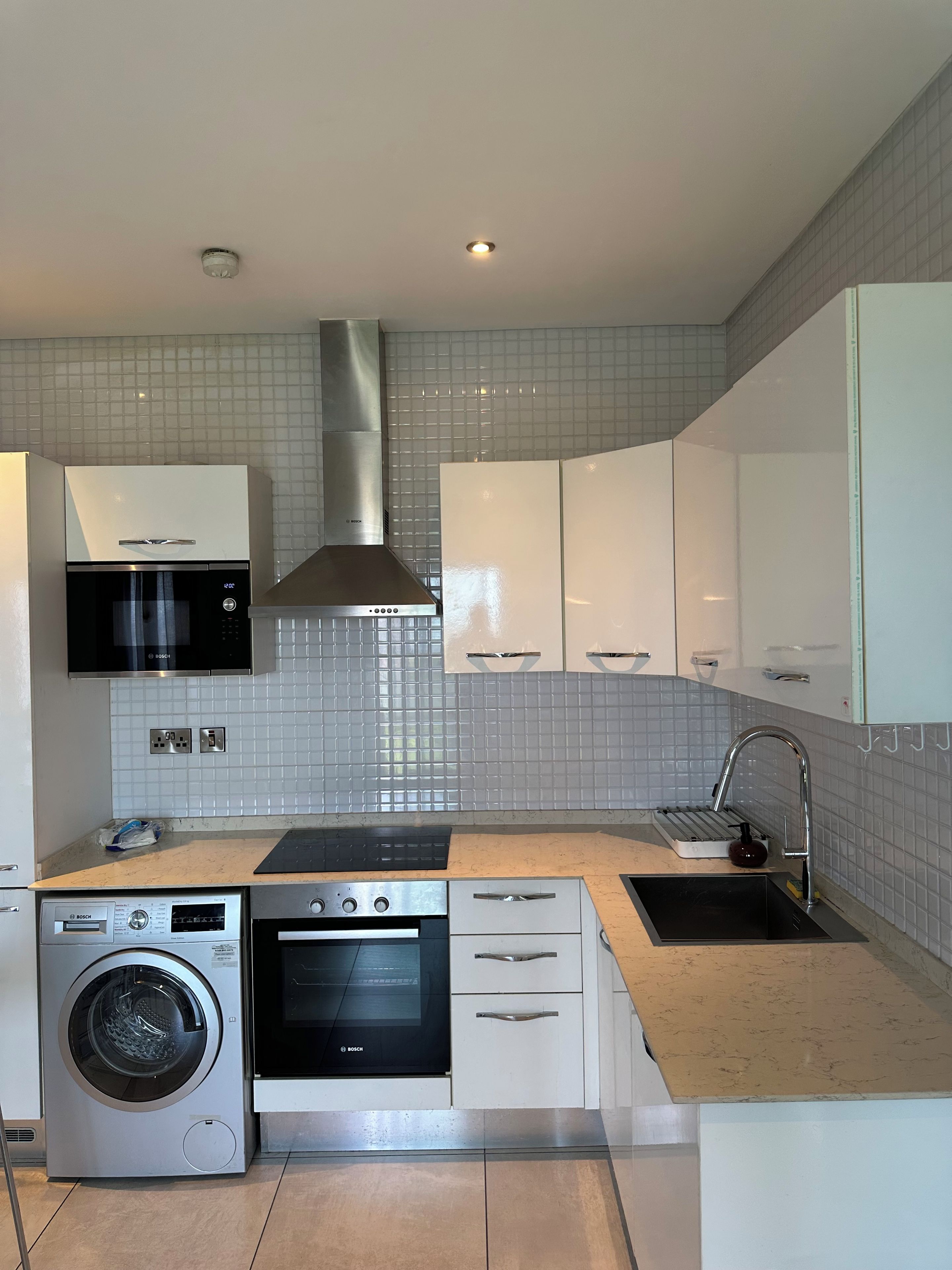 A modern kitchen with Bosch appliances, including a microwave, oven, and washing machine, with white cabinets and a tiled backsplash.