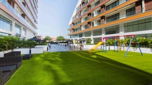 Ein modernes Gebäude mit mehreren Etagen, einem Innenhof mit künstlichem Rasen und einem Kinderspielplatz mit Schaukeln und Rutsche.