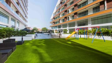 Um edifício moderno com vários andares, um pátio com grama sintética e uma área de recreação infantil com baloiços e escorrega.
