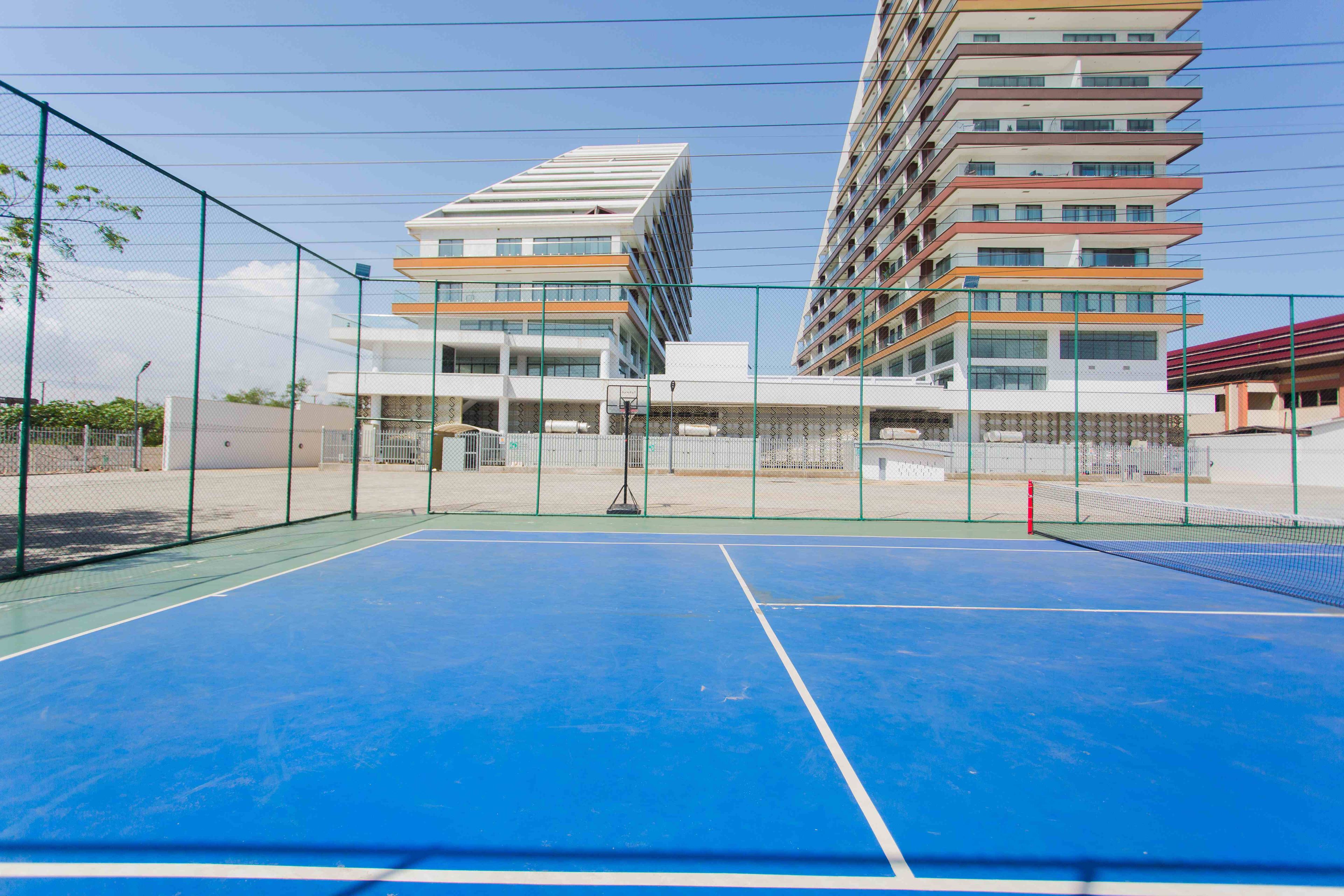 Una cancha de tenis exterior con red, rodeada por una cerca de malla metálica, y adyacente a un edificio de varios pisos con balcones.