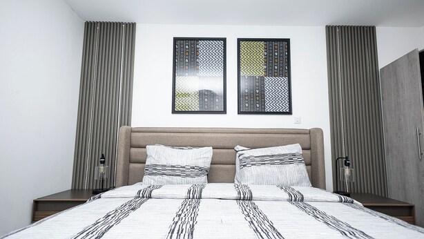 A modern bedroom with a large bed, two nightstands, a headboard, and two framed wall art pieces. - Peravi at The Signature (Accra)