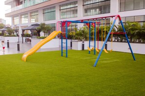 Um parquinho ao ar livre com escorrega e balanços, cercado por um gramado bem cuidado e um edifício ao fundo.