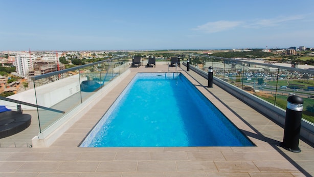 Ein Pool auf dem Dach mit Glasgeländer, Liegestühlen und Blick auf die Stadt.