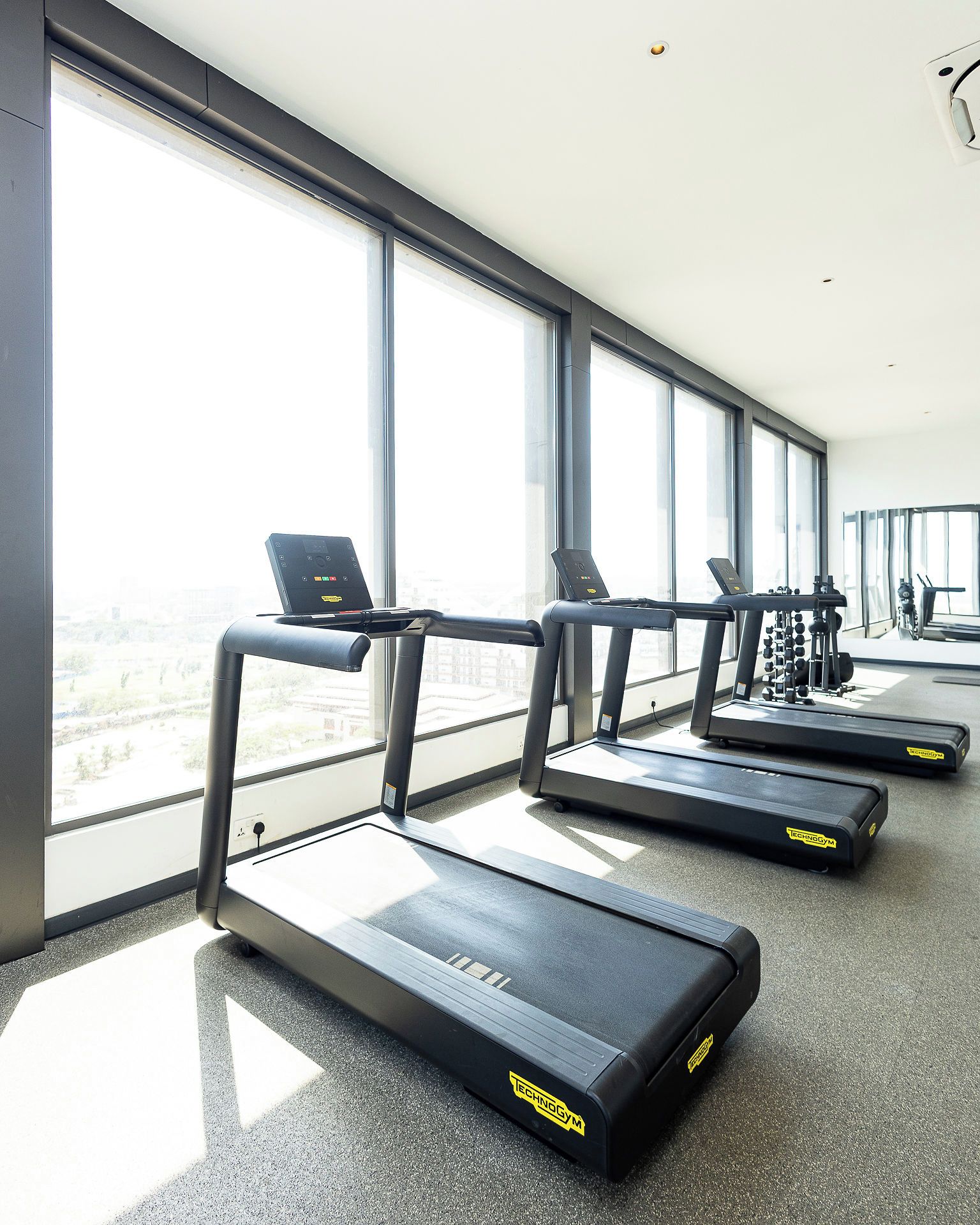 Un gimnasio con cintas de correr y equipo de ejercicio, ventanas amplias y vista a la ciudad.