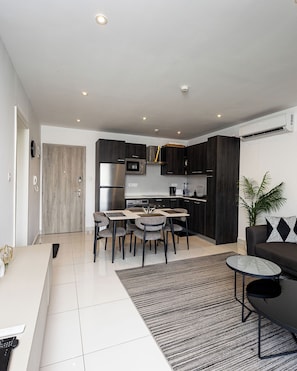 A modern kitchen with dark cabinetry, a stainless steel refrigerator, and a microwave. There is a dining area with a table and chairs. - Peravi at The Signature (Accra)