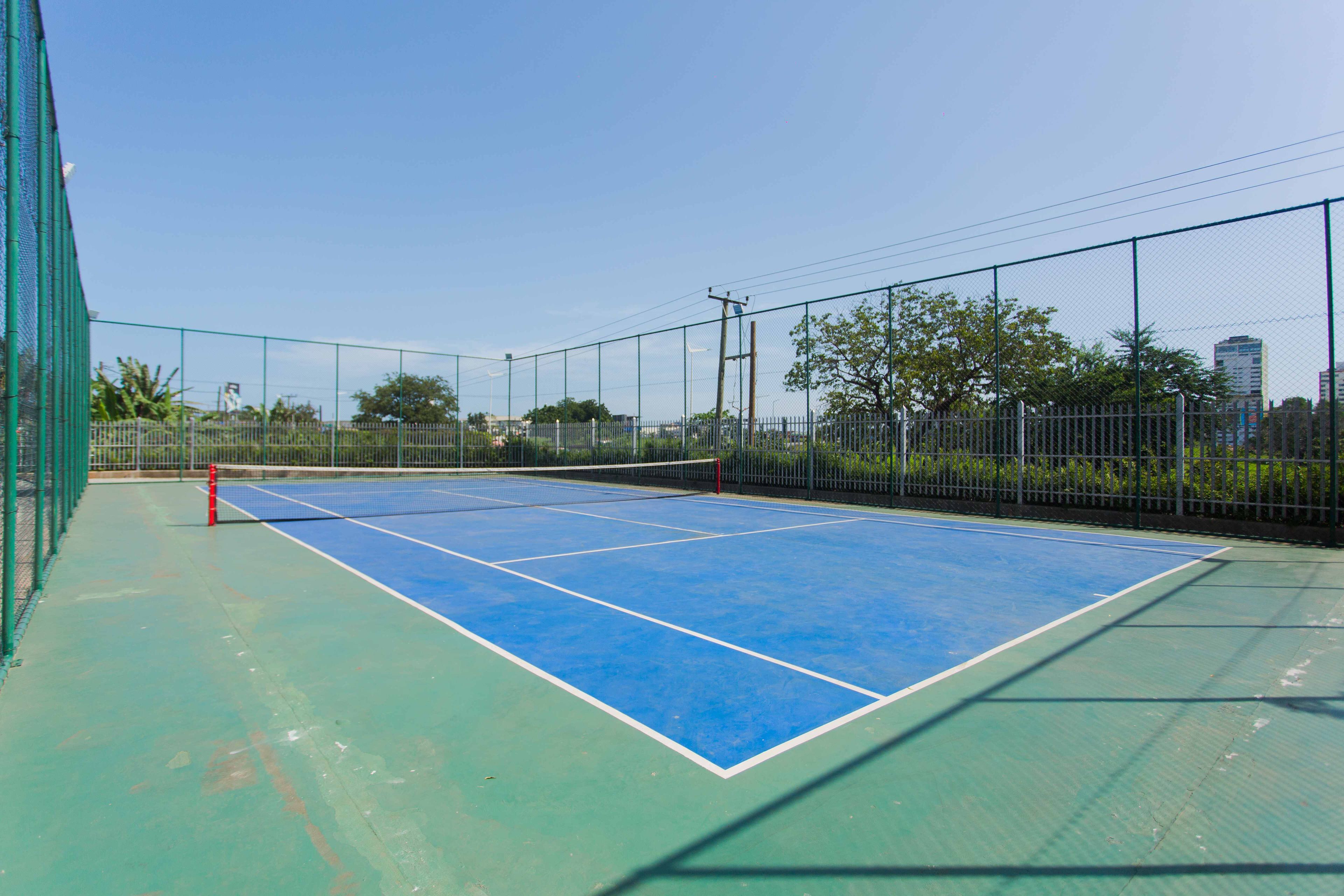 An outdoor tennis court with a net, surrounded by a chain-link fence and trees.