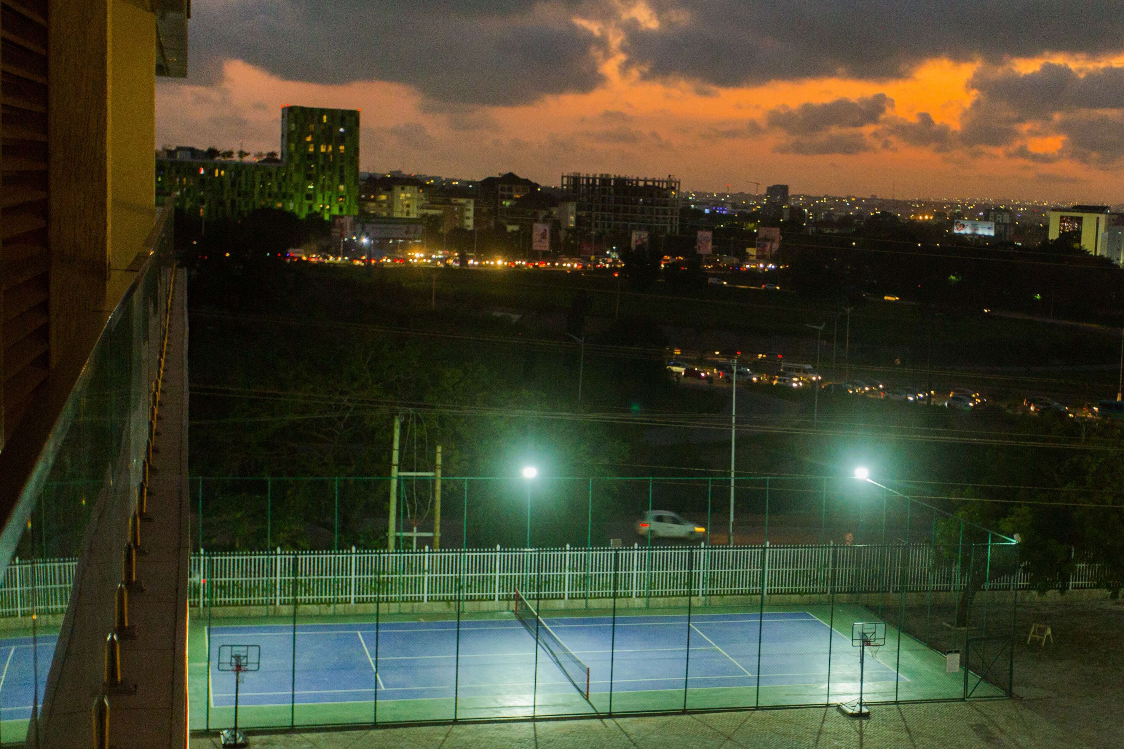 An outdoor basketball court with a fence, a basketball hoop, and a view of a cityscape at dusk.
