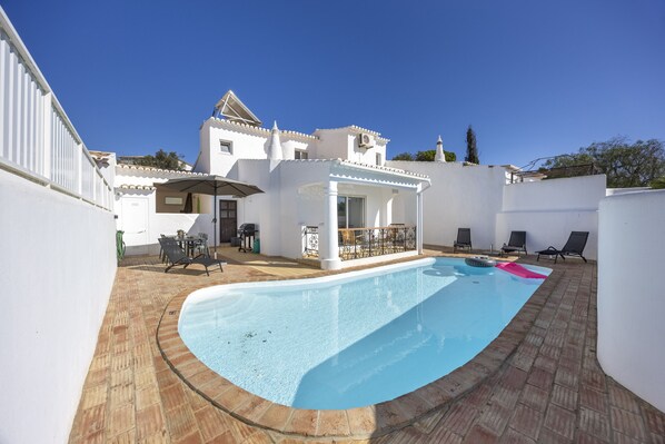 A swimming pool with a tiled deck, a patio area with outdoor furniture, and a building with a tiled roof and white walls.