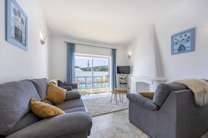 A living room with a grey sofa, a smaller grey armchair, a TV, and a fireplace. There is a balcony with a pool and outdoor furniture. - Casa Lília | Mte S Pedro 2 (Luz)