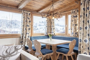 Ein gemütliches Zimmer mit Holzbalkendecke, einem runden Tisch mit blau gepolsterten Stühlen und einer Bank mit floralen Vorhängen.