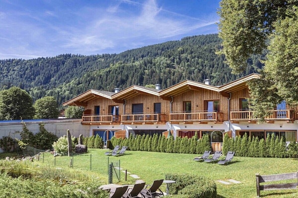 Un bâtiment de style chalet doté de nombreux balcons, d’un jardin bien entretenu avec des chaises longues et d’une montagne boisée en arrière-plan.