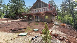 A two-story wooden cabin with a balcony, surrounded by a natural landscape with trees and a dirt ground. - Pine Cabin w View, Fireplace, Deck & Game Room (Pine)