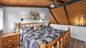 A bedroom with a wooden ceiling, a bed with a plaid comforter, two wall-mounted lamps, and a deer painting. - Pine Cabin w View, Fireplace, Deck & Game Room (Pine)