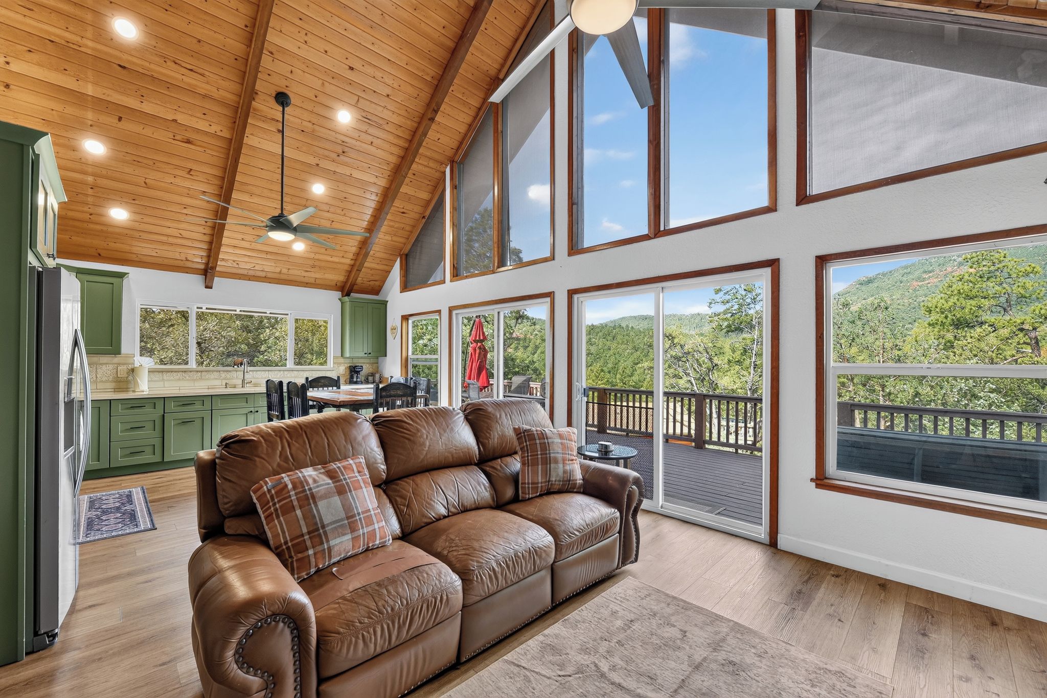 A spacious living room with a leather sofa, a dining area, and large windows offering a view of a forested landscape.