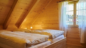 Una stanza in legno con soffitto spiovente, un letto con lenzuola bianche e una finestra con tende in pizzo.