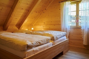 Een kamer met houten wanden en een schuine kap, een bed met witte lakens en een raam met kantgordijnen.