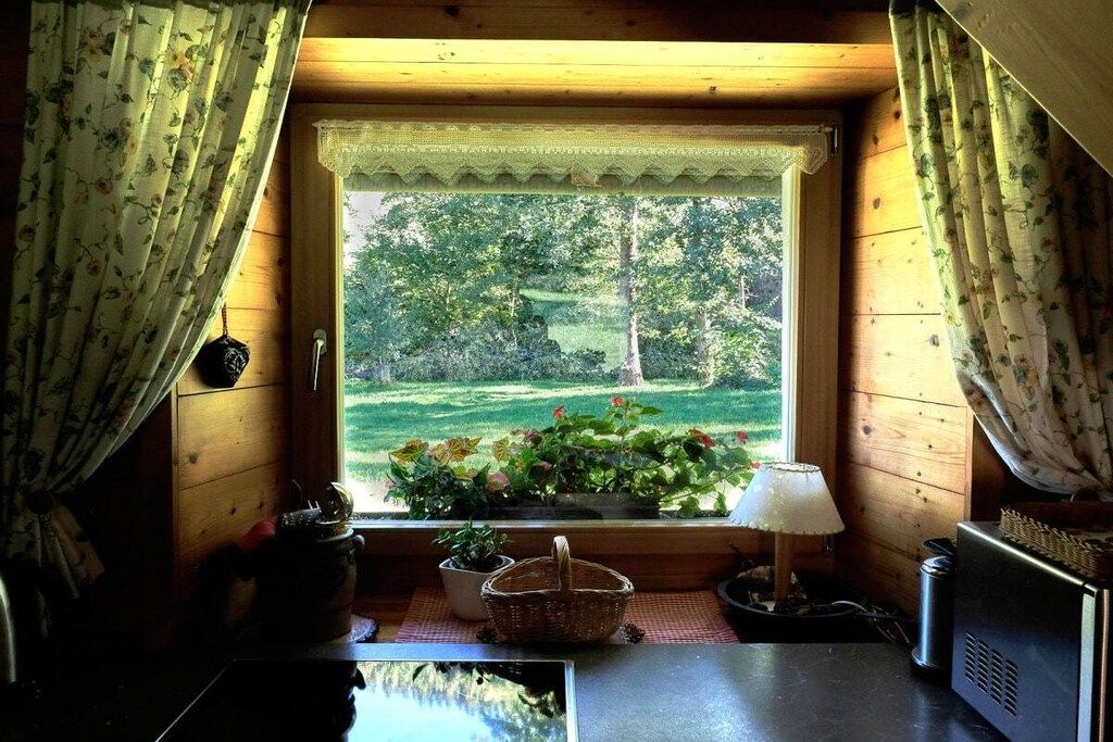 A cabin interior with a window overlooking a garden, a table with a lamp, and a radio.