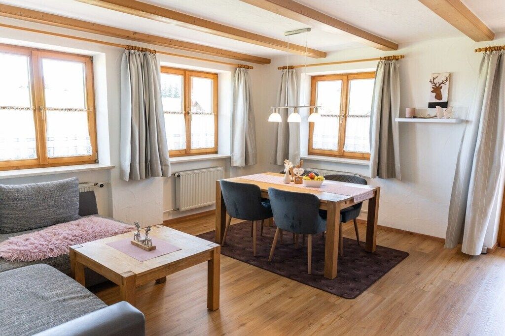 Un salon confortable avec un canapé, une table basse et une table à manger avec des chaises. De grandes fenêtres sont agrémentées de rideaux, il y a un radiateur et une étagère où sont disposés des objets décoratifs.