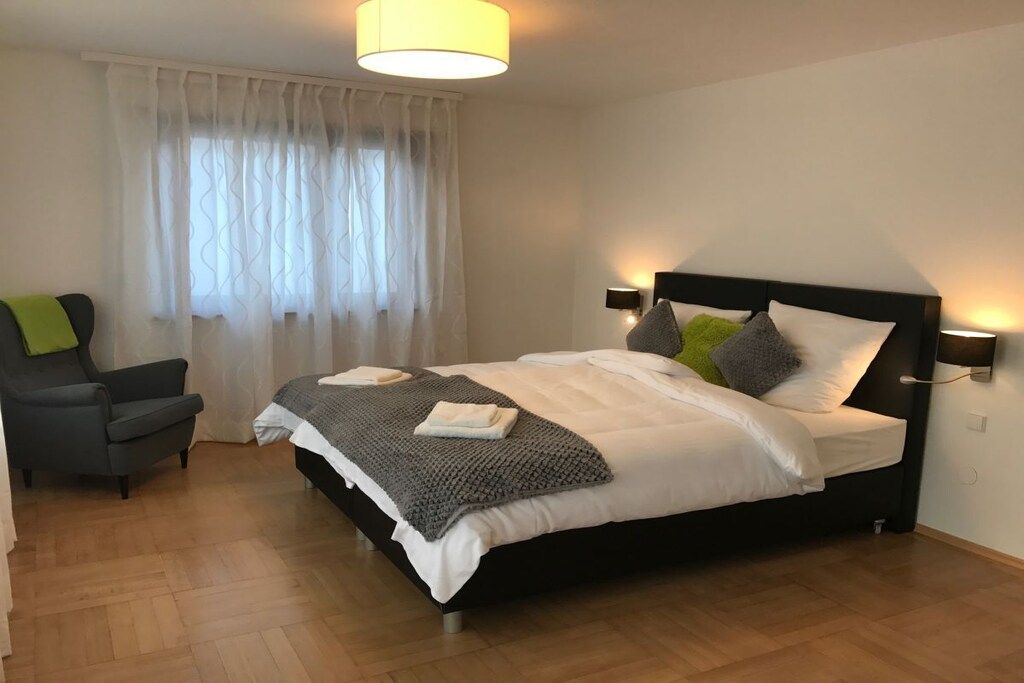 Un dormitorio con una cama, una silla, un ventanal con cortinas y una lámpara fijada a la pared.