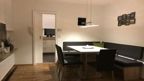 Ett modernt matområde med ett vitt bord, svarta stolar och en bänk. Genom en öppen dörr kan man se in i köket.