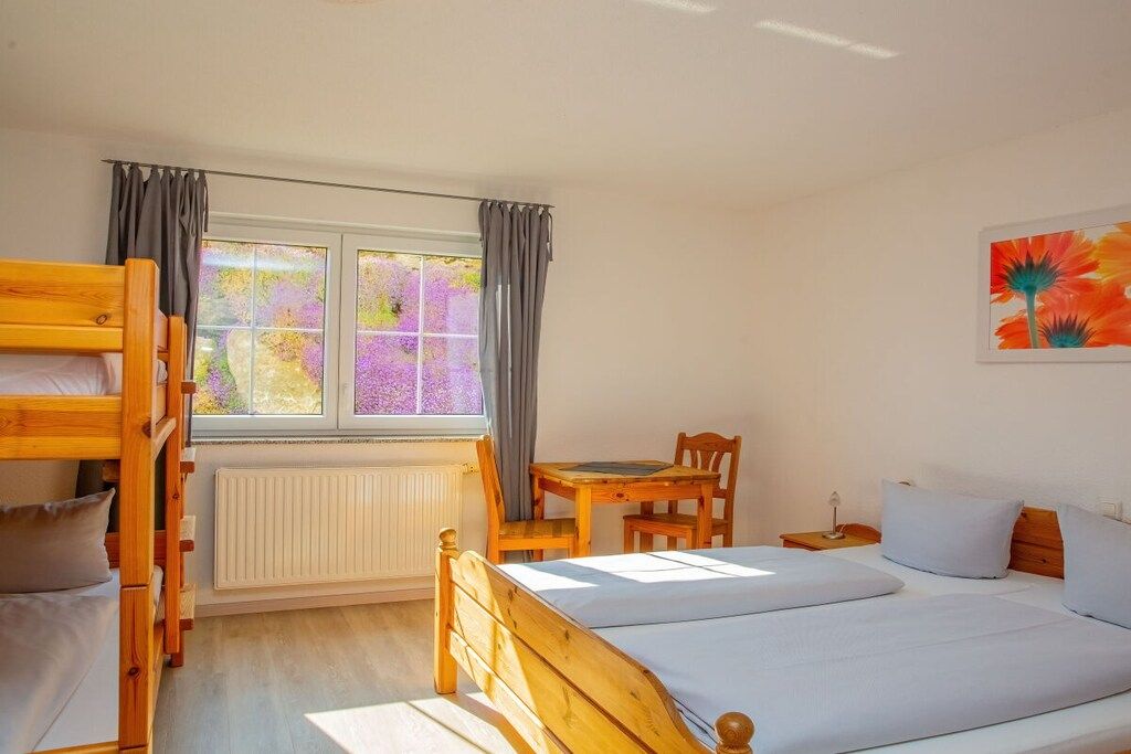 Une chambre avec un lit superposé, un lit simple, une table avec des chaises, un radiateur et une fenêtre avec des rideaux.