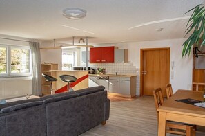 A modern living room with a grey sofa, a dining table with chairs, a kitchen area with red cabinets, and a wooden door. - Holiday apartment with 1 bedroom (Meersburg)