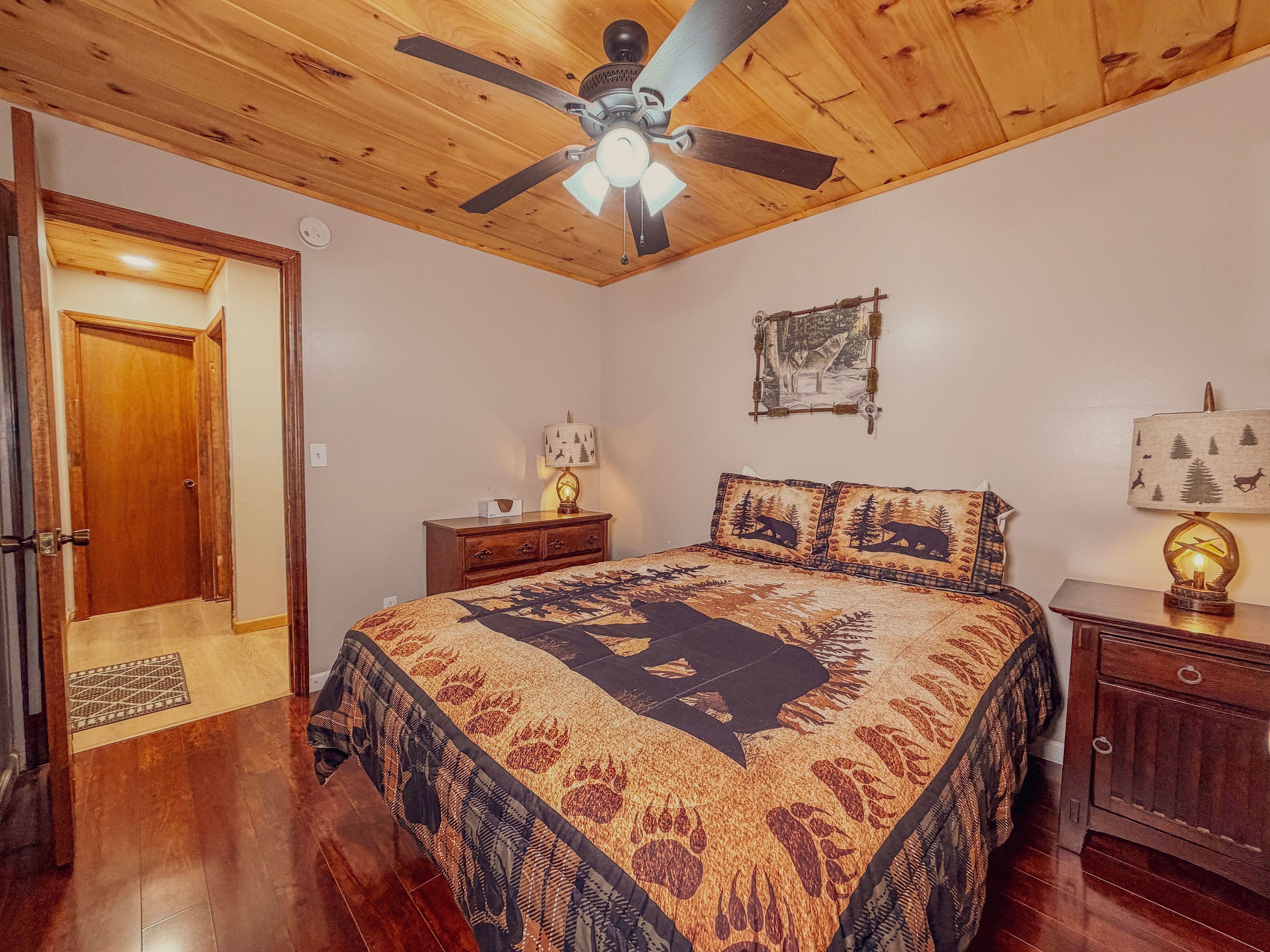 A bedroom with a wooden ceiling, a bed with a patterned bedspread, a nightstand with a lamp, a wall clock, and a door.