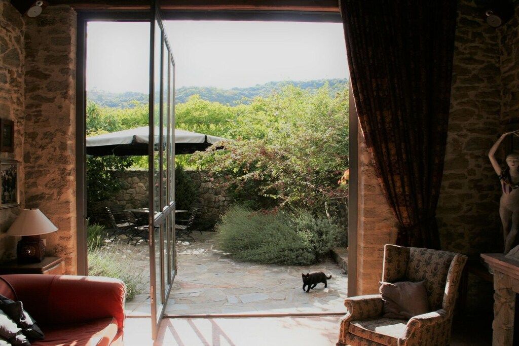 A room with a stone wall, a red sofa, a patterned armchair, and a view of a garden through a glass door.