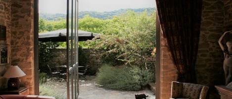 A room with a stone wall, a red sofa, a patterned armchair, and a view of a garden through a glass door.