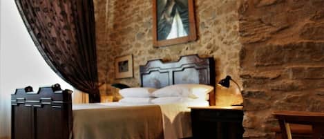 A bedroom with a stone wall, a bed with a wooden headboard, a nightstand, and a lamp.